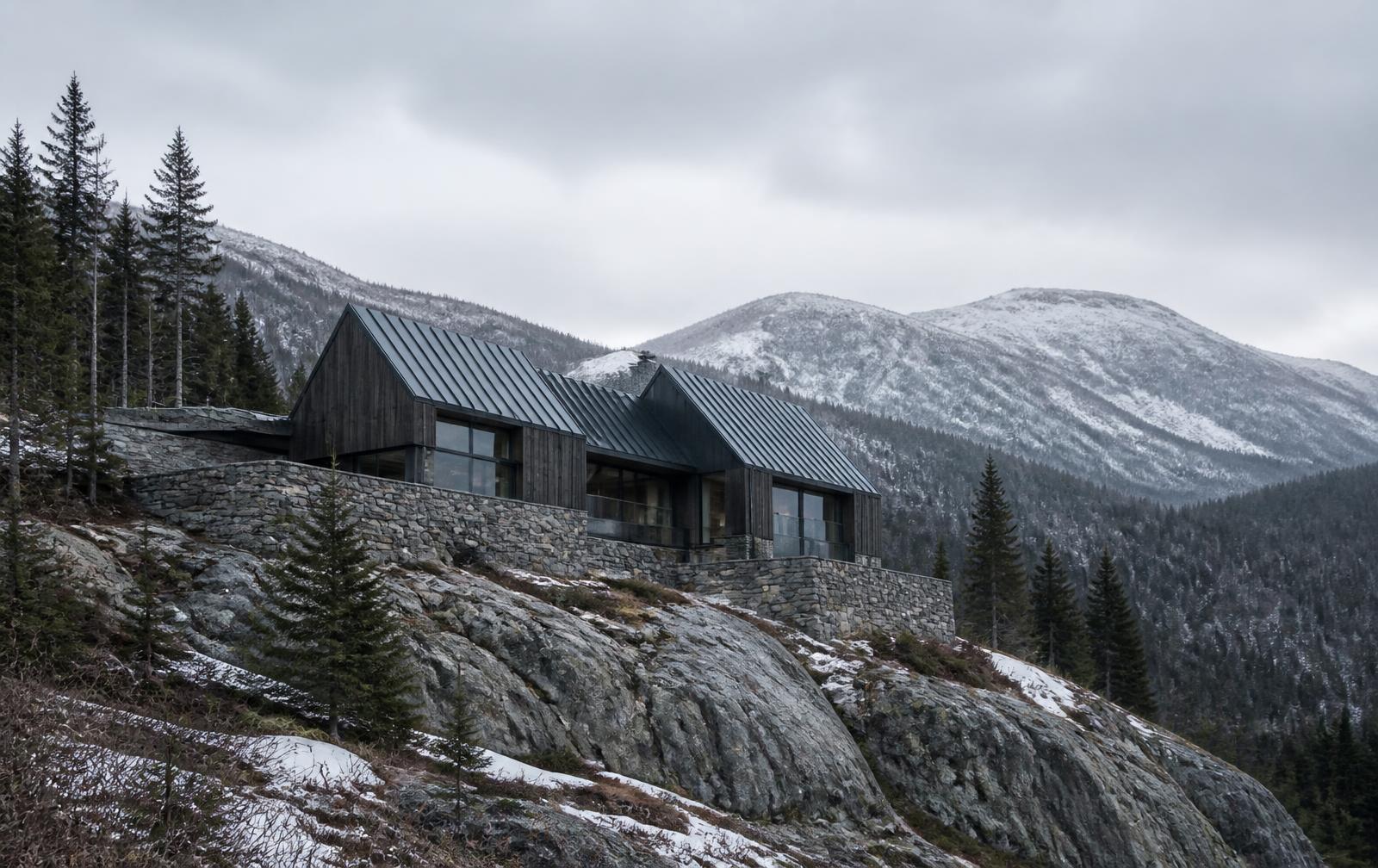 Concept visualization of a mountain residence seen from below across rocky terrain, its granite base walls stepping with the topography