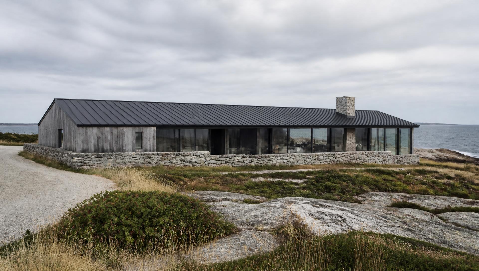 Concept visualization of the approach to the residence — one long low cedar volume under a dark standing-seam metal roof on a granite base wall, seen across ledge and beach grass with the Atlantic beyond