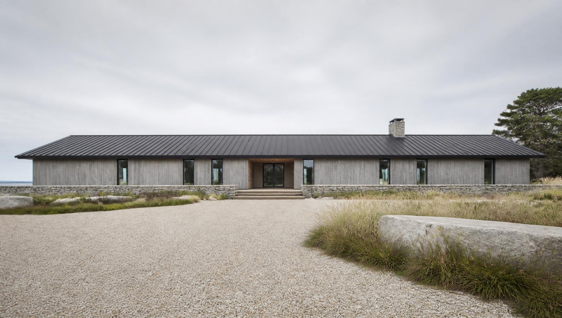 Concept visualization of the land-side entry elevation — a single long low volume in weathered vertical cedar on a dry-stack granite base wall, under a shallow-pitch dark standing-seam metal roof with a recessed white oak entry