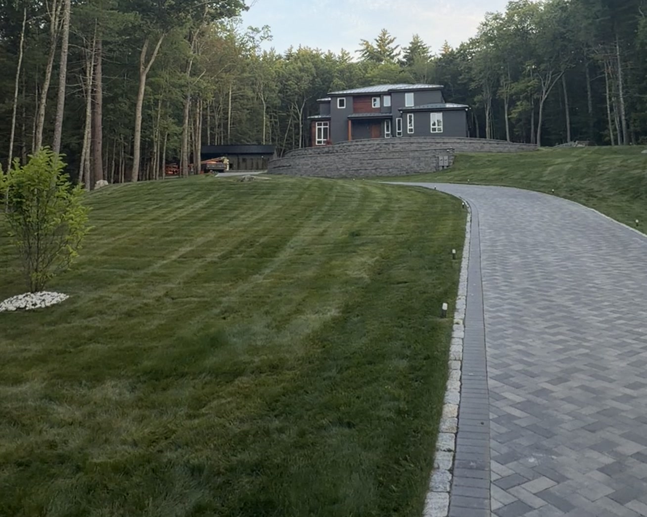 Paved driveway curving up a graded lawn to the Bedford Residence, a dark-clad house with standing-seam metal roof above a curved stone retaining wall
