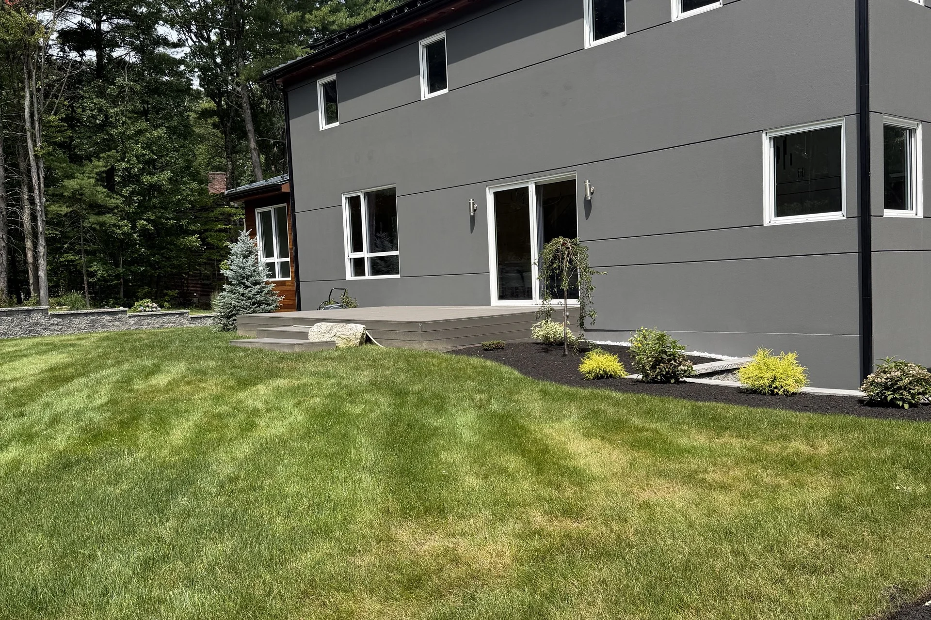 Rear elevation of the Bedford Residence with charcoal siding, white-trimmed glazing and a graded lawn edged in dark mulch beds