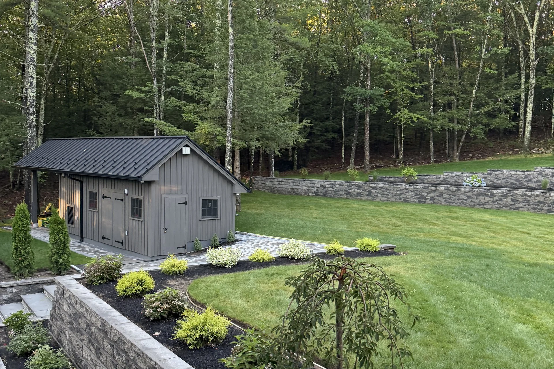 Grounds at the Bedford Residence with tiered stone retaining walls, planting beds and a matching outbuilding at the woodland edge
