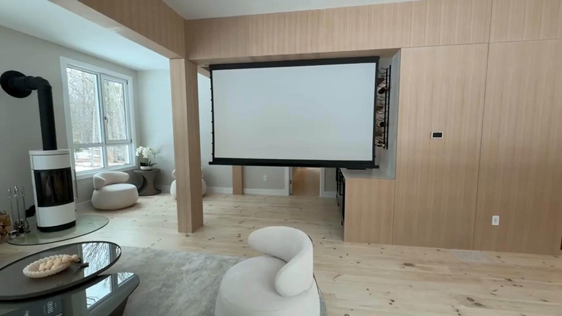Living room at the Bedford Residence with the projection screen lowered from the white oak portal wall, a freestanding stove and wide plank flooring